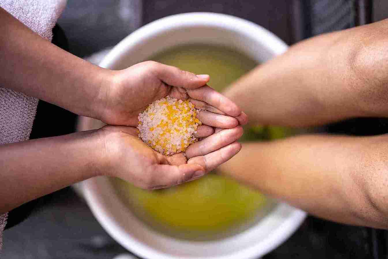 FloWü - Salzfußbad Füße stehen in einer gefüllten Wasserschüssel. Zwei Hände halten Salz darüber
