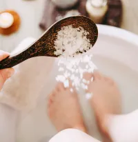 FloWü - Zubereitung Solefußbad Zwei Füße in Schüssel mit Wasser. Im Vordergrund Solelöffel mit langsam herausgeschüttetes Salz